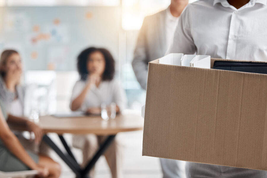 Employee leaving office with a box after being laid off with coworkers in the background
