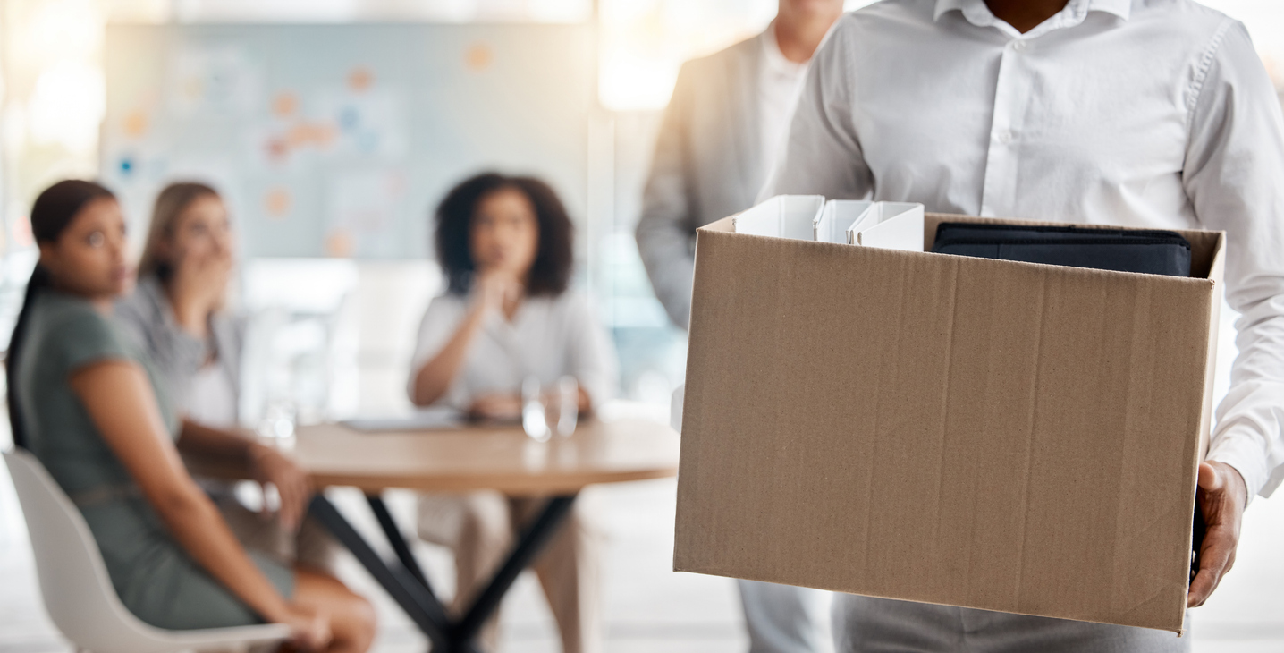 Employee leaving office with a box after being laid off with coworkers in the background